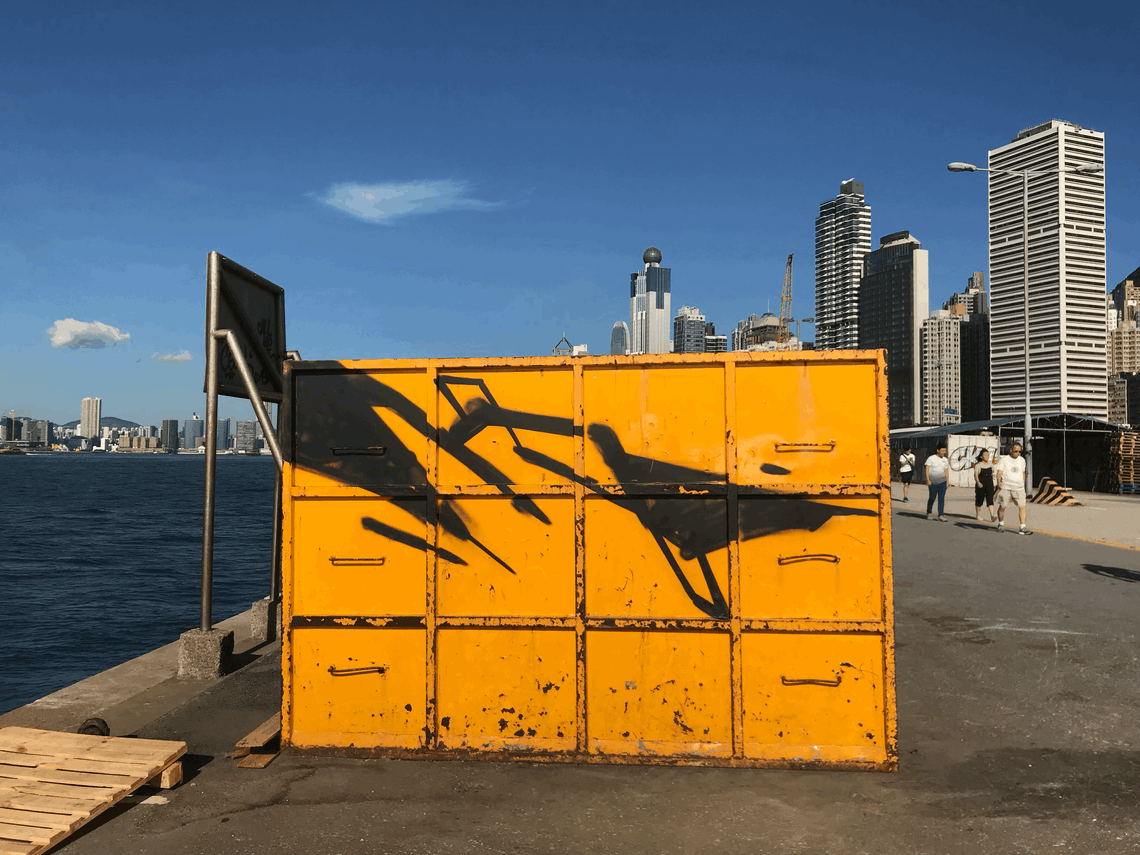 Black spray paint on a bright yellow container on a pier. Blue sky and tall buildings in the background.