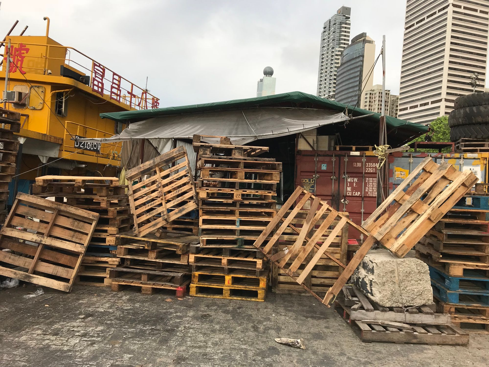 Wooden pallets strewn in front of a make shift shelter on a pier. A tug boat is on the left. 