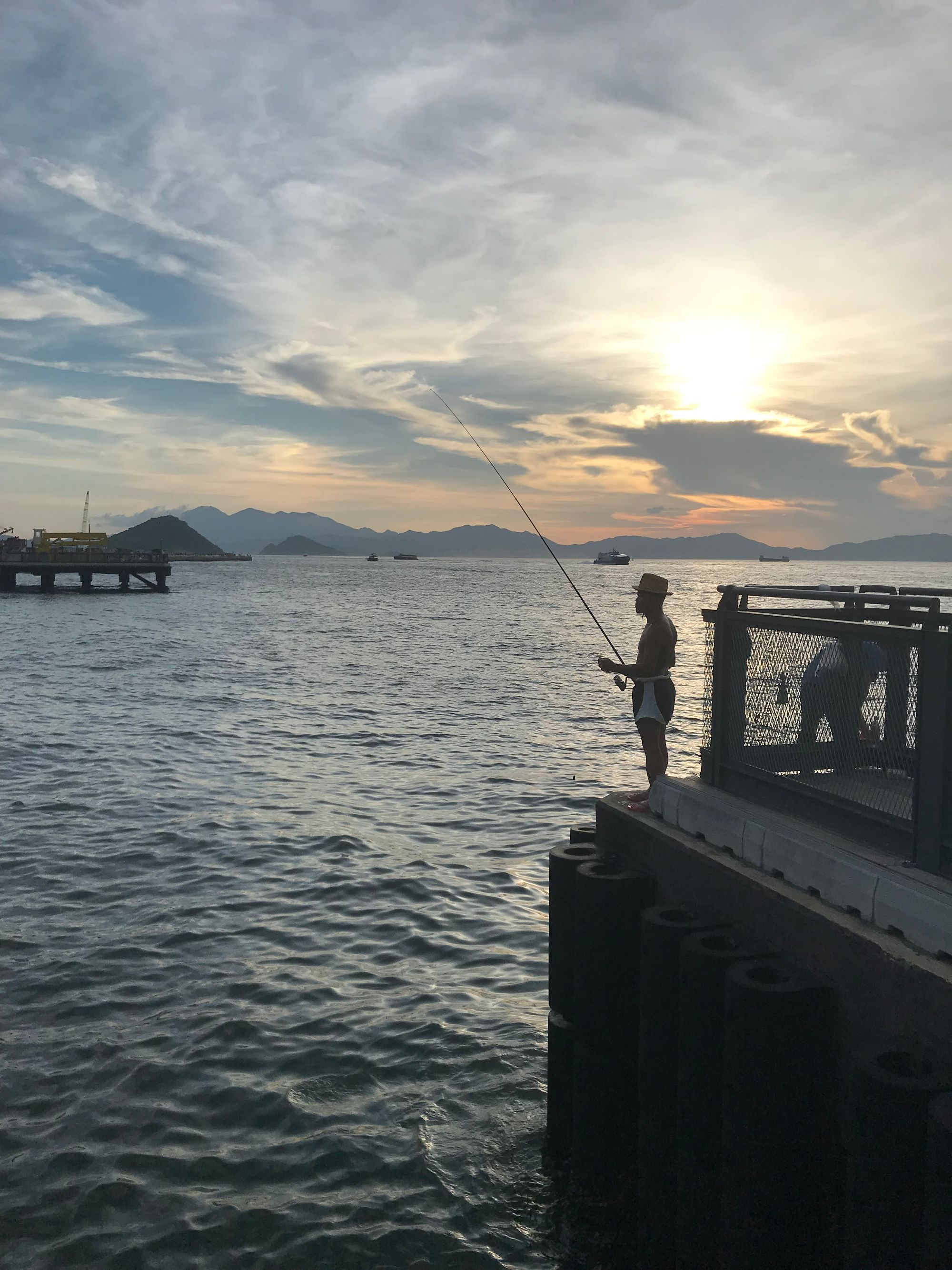 A shirtless man fishes with a rod on the edge of a pier with the sun beginning to go down behind him