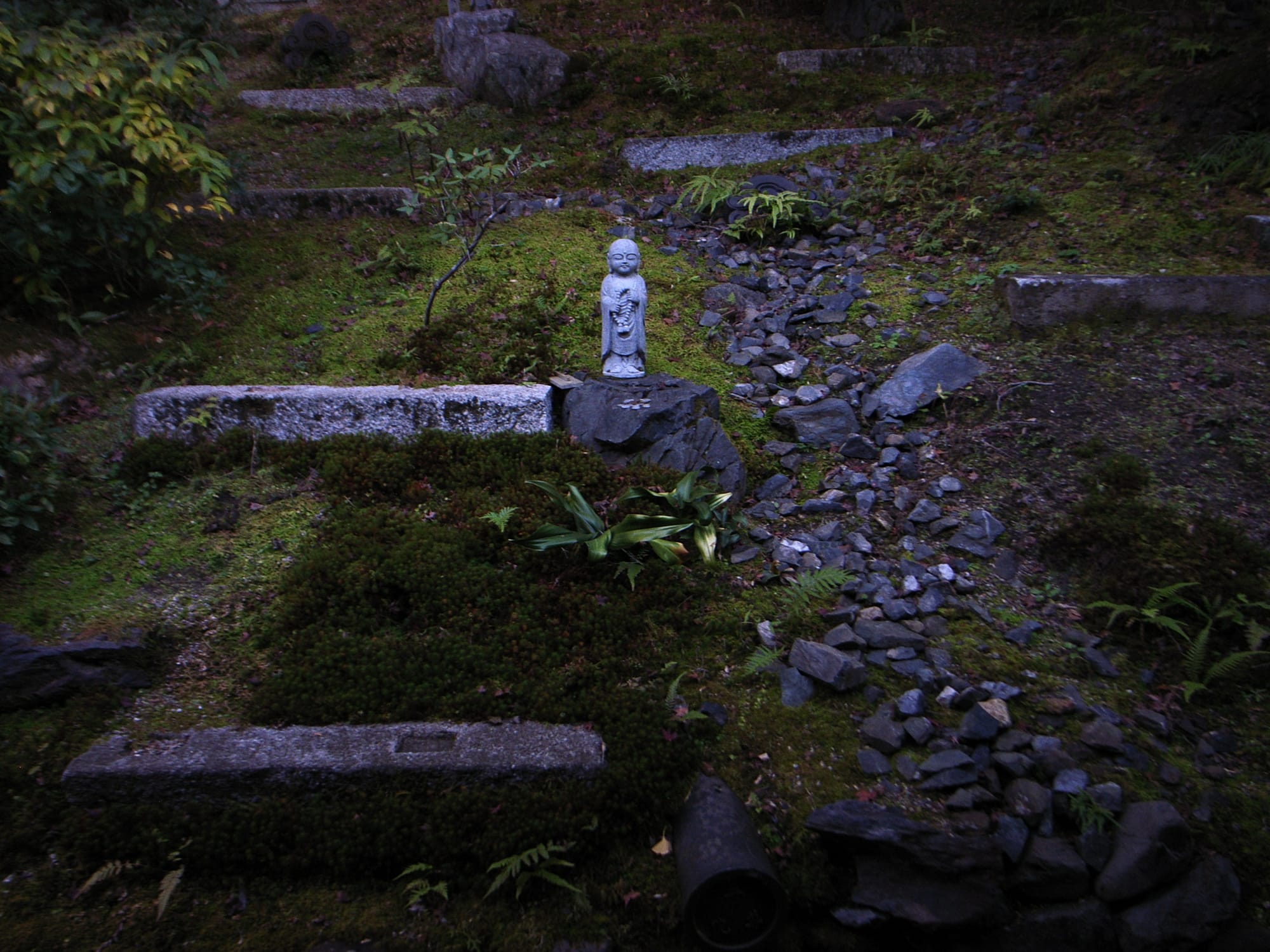 A little stone Bodhisattva amid stone slabs, rocks, grass, and moss.