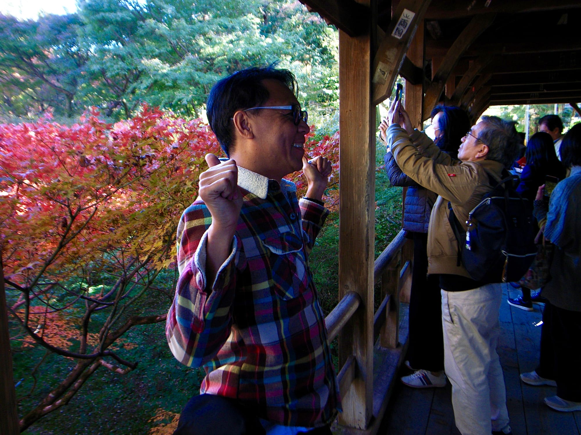 A man in a plaid jacket poses with his thumbs up in the foreground. Another man slight further back raises his phone to take a picture.