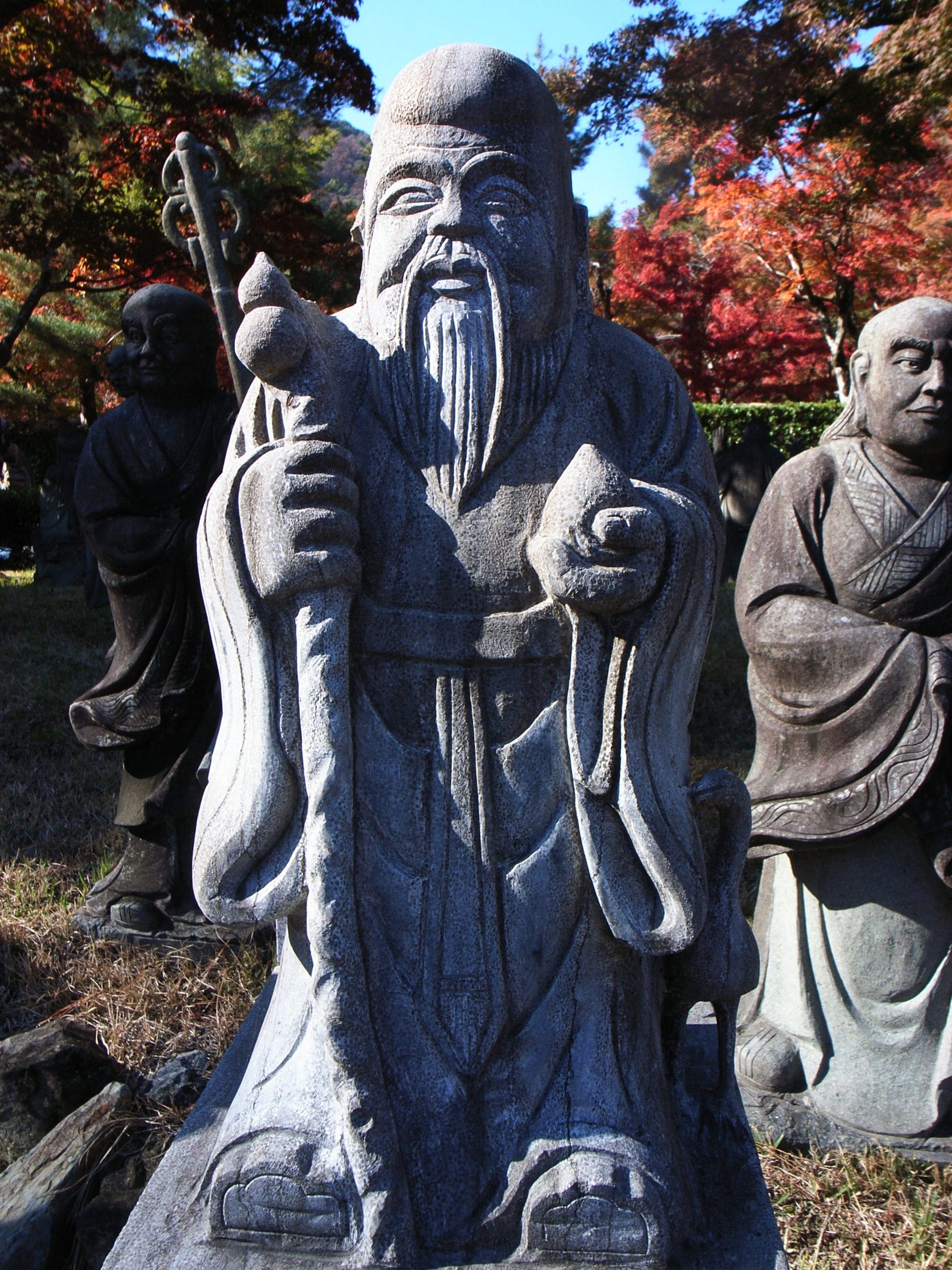 A stone statue of an arhat, one of Buddha's followers, with some red autumn foliage behind it.