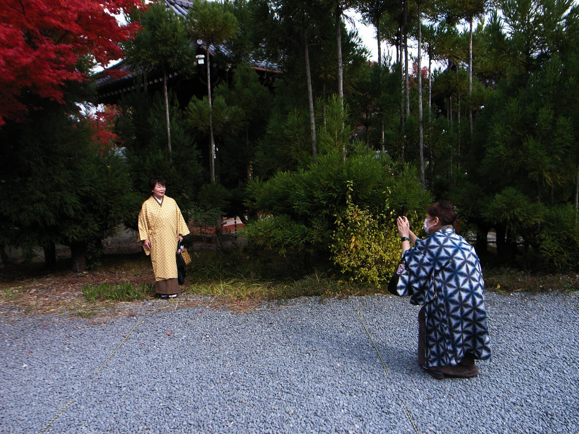 A woman in a blue-patterned coat takes a picture of another woman in a kimono with some red foliage above.