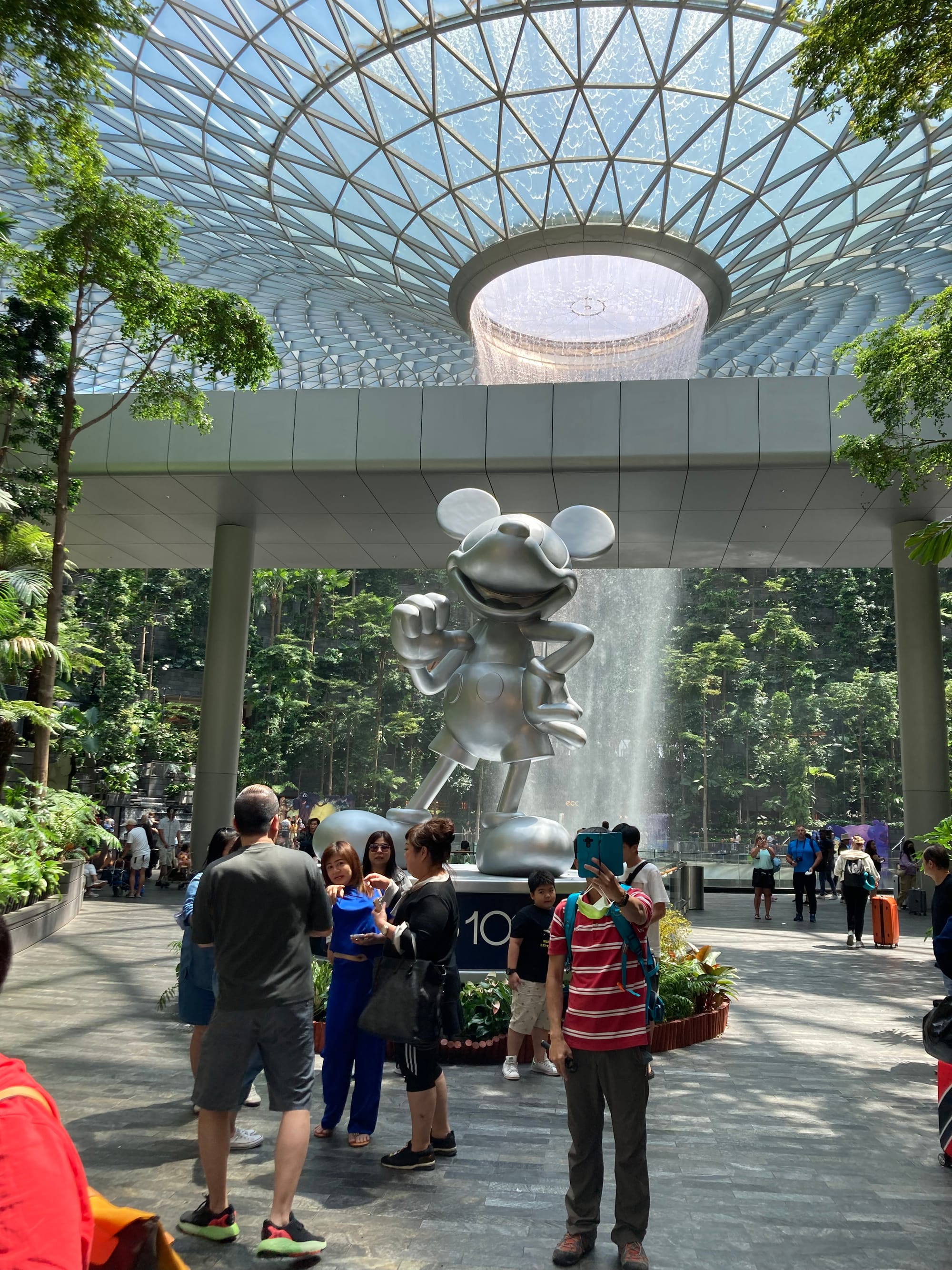 Visitors pose for a photo before a platinum Mickey Mouse statue. The indoor waterfall is in the background.