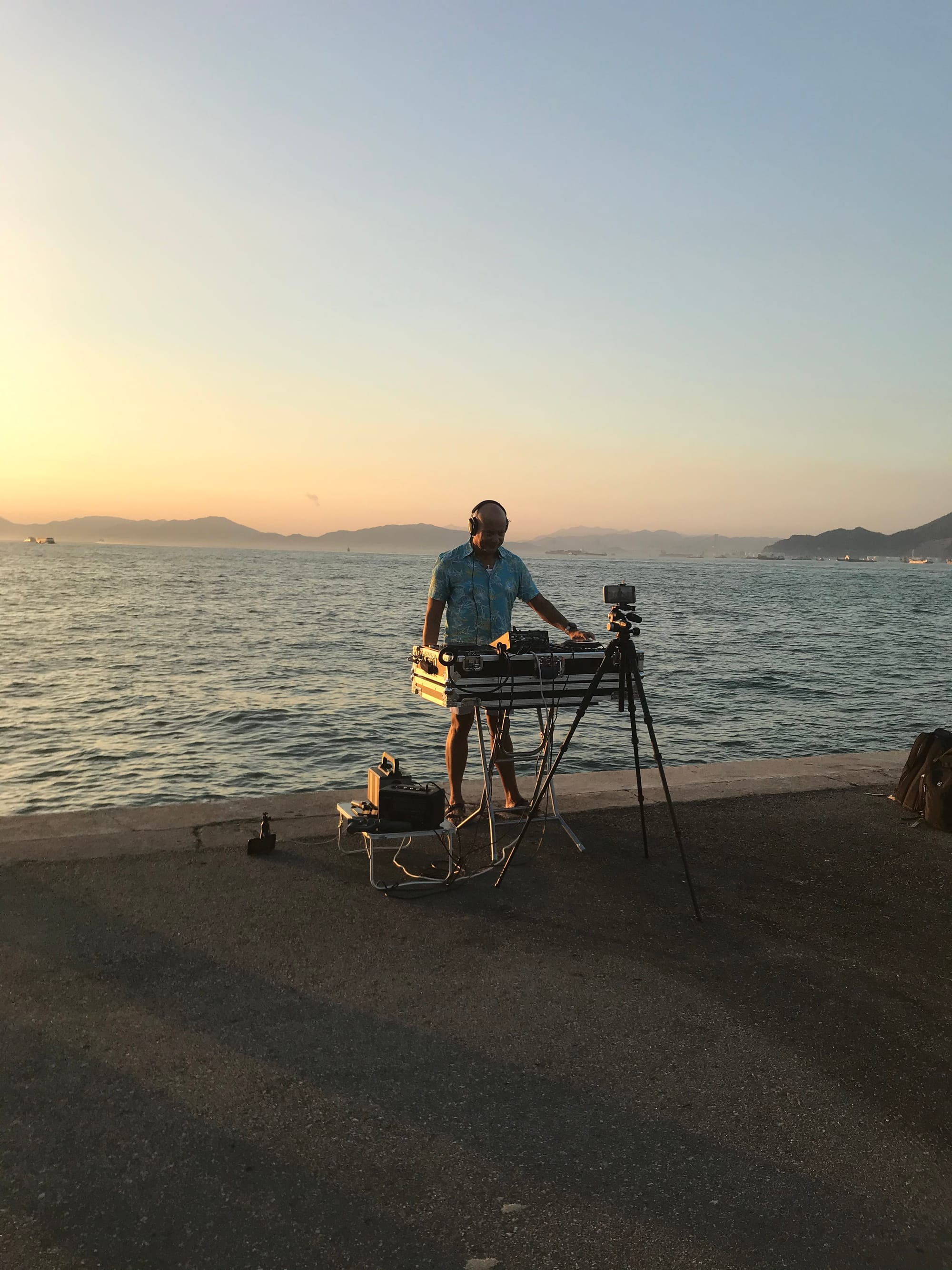 A man playing a DJ set on the pier. There is a camera on a tripod filming him and the sea and islands bathed in the pink glow of the setting sun are behind him.
