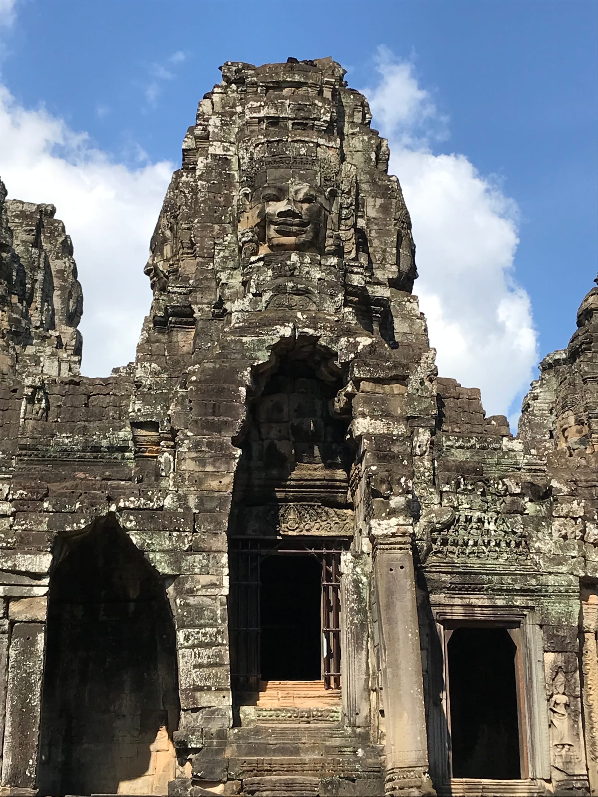 A smiling stone Buddha face is embedded in a stone tower. There are doorways beneath the tower.