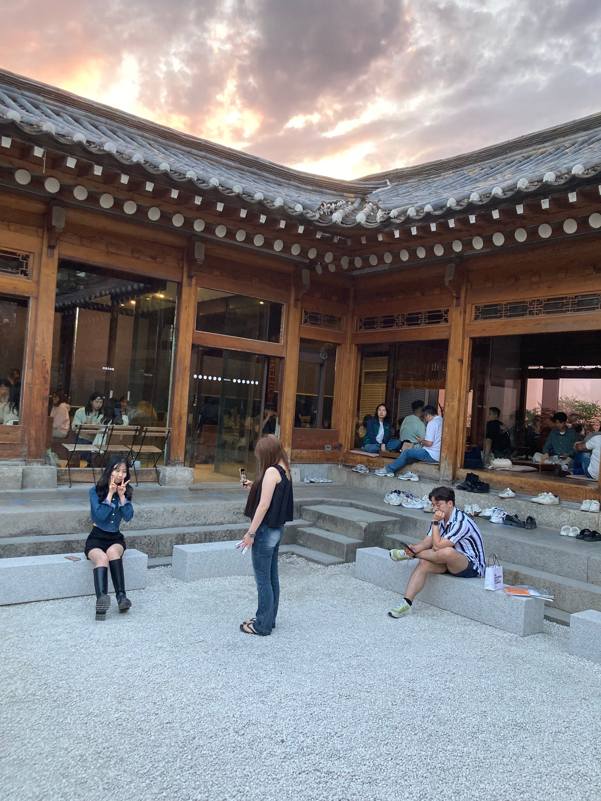 A traditional wooden house serves as a busy cafe. In front of it, two girls are taking a photo and a man is checking his phone.