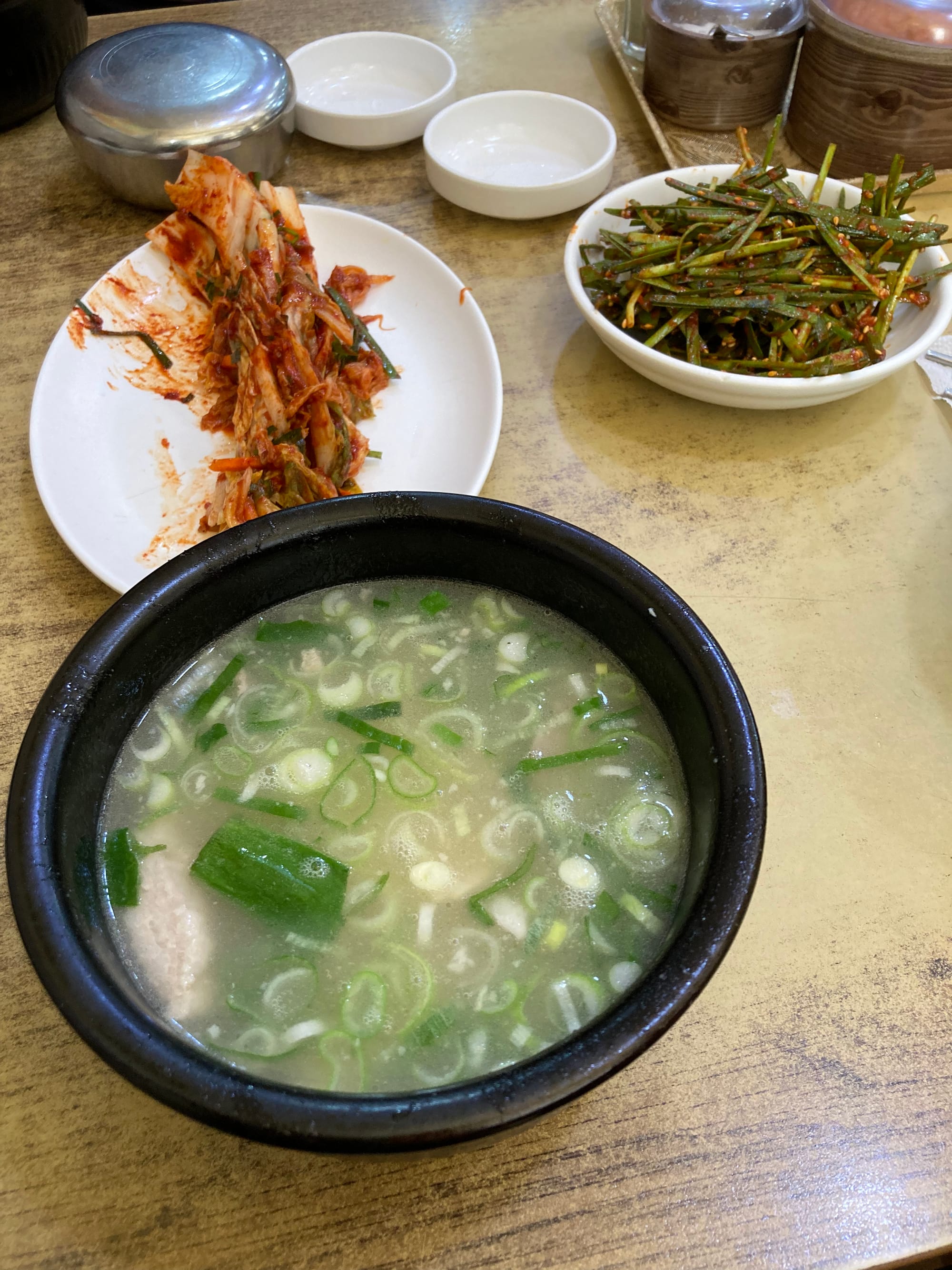 A bowl of pork broth with spring onion. Plates of cabbage kimchi and chives kimchi are in the background.