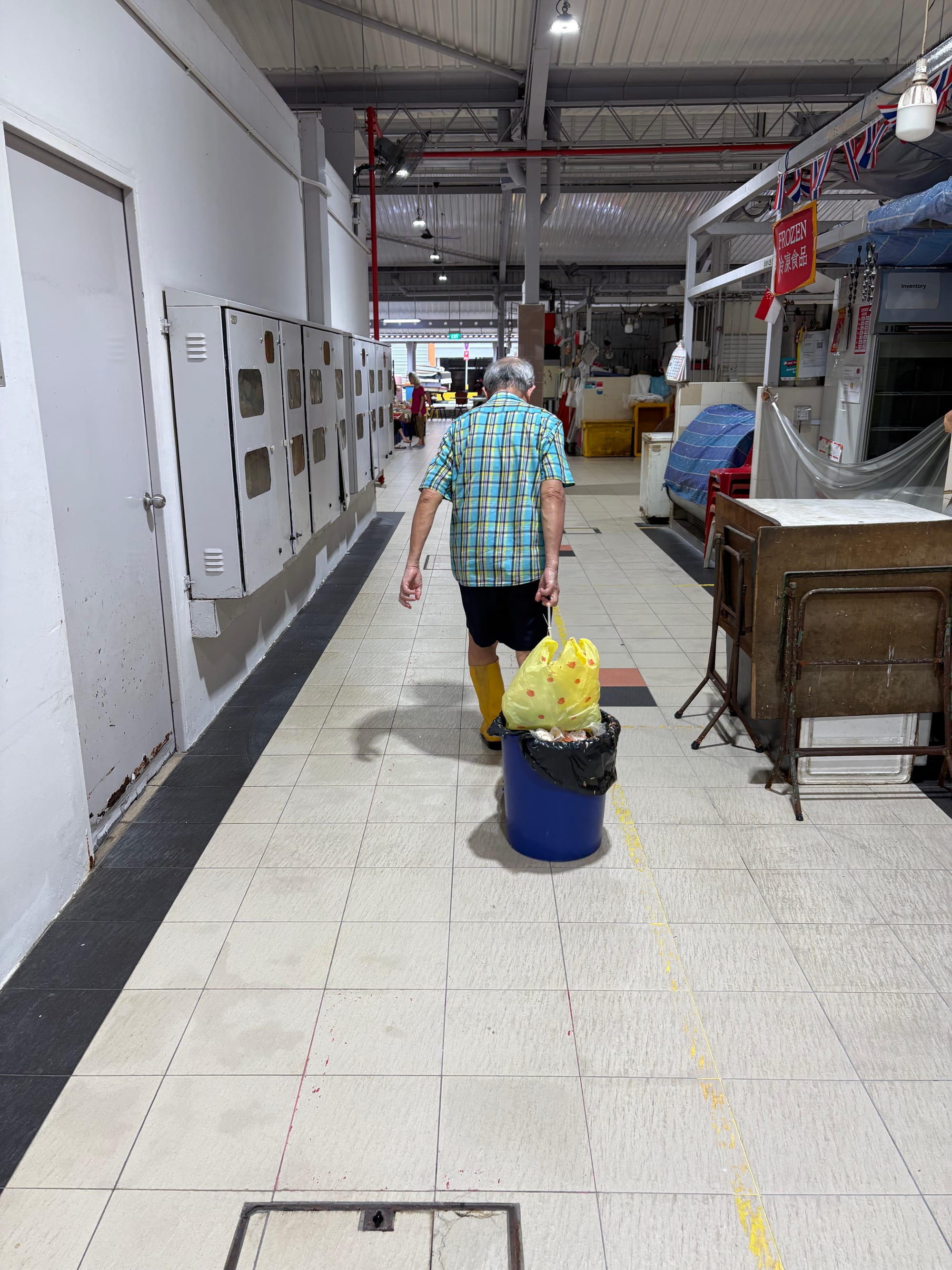 An old man drags a blue plastic bin with a yellow bag on it through the market.