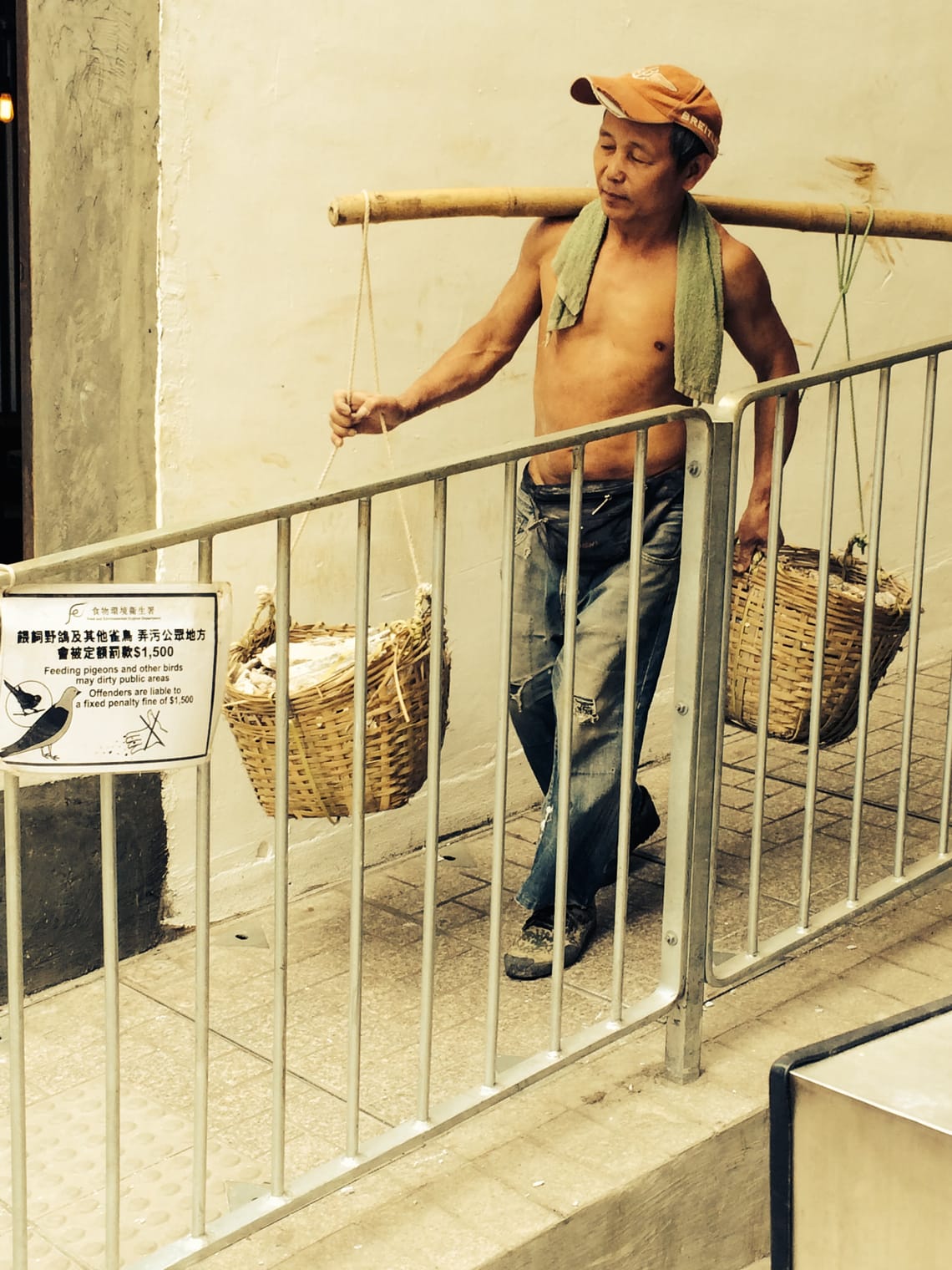 A shirtless man wearing a cap carries a bamboo pole with two woven baskets attached to it.